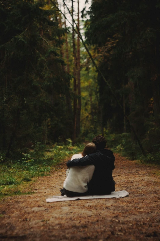 two people sit on the ground together and look out at the woods