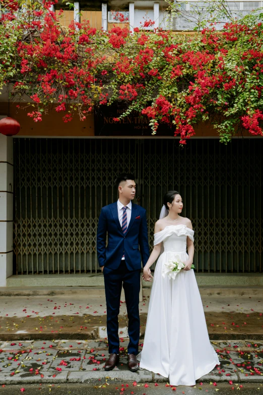 a man and woman posing in front of a building with red flowers