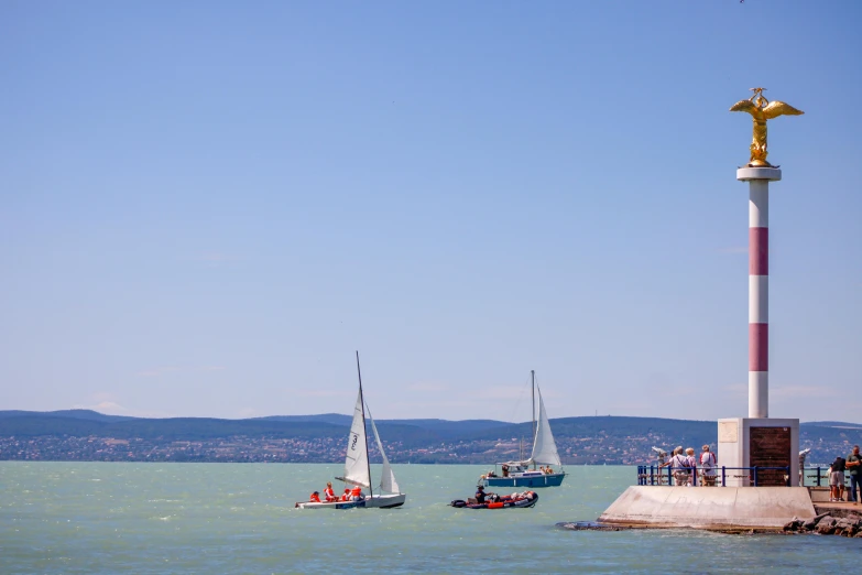 sailboats passing on the water with a lighthouse and a statue in the background