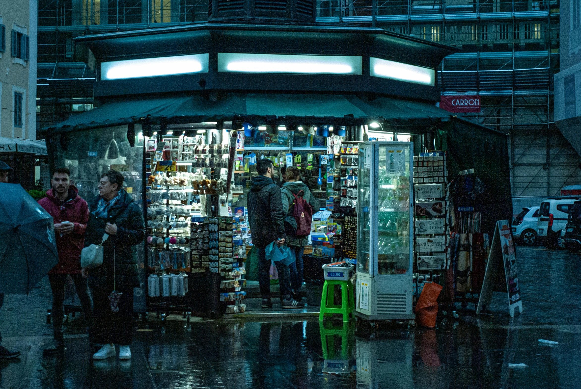 people are standing outside of a convenience store in the rain