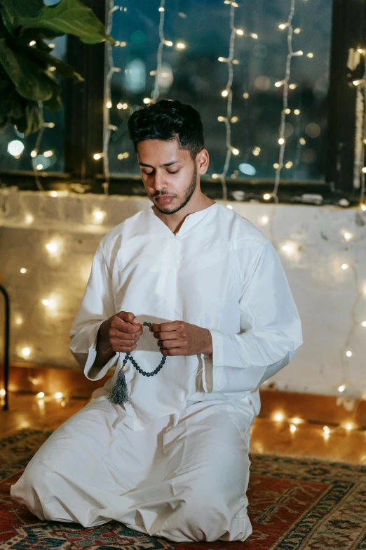 a man kneeling down holding a rosary and wearing white clothing
