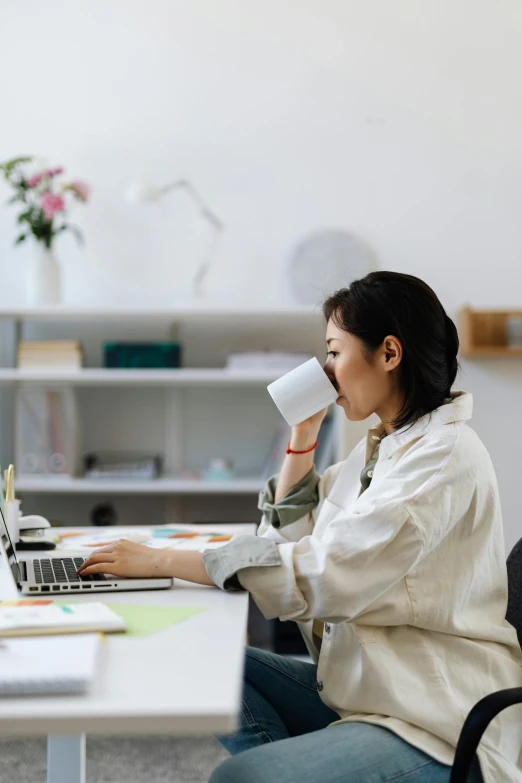 a woman is drinking from a mug while using her laptop