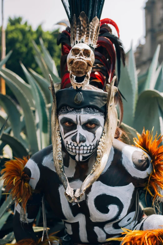 a masked person with painted skin, white face paint and silver body paint, with feathers in his hair