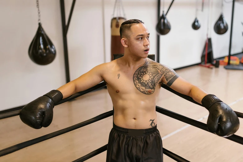 young man with tattoos wearing boxing gloves leaning on ropes in a gym