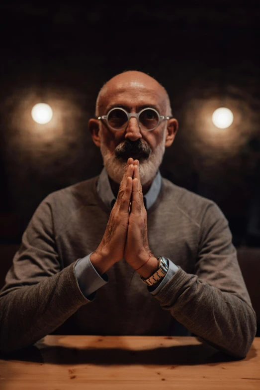 a man wearing glasses sitting at a table with his hands clasped to his face