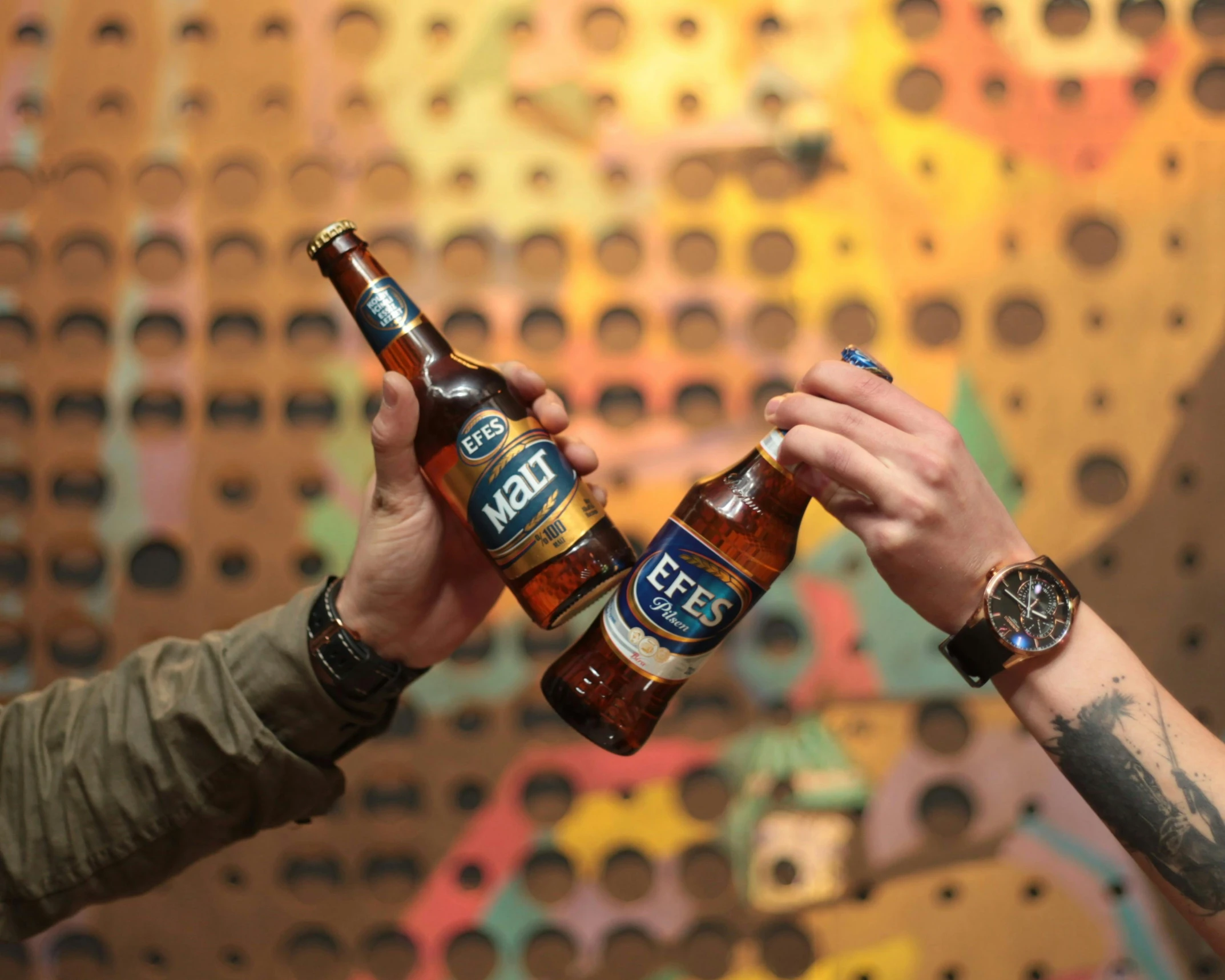 two hands holding beer bottles in front of an array of designs