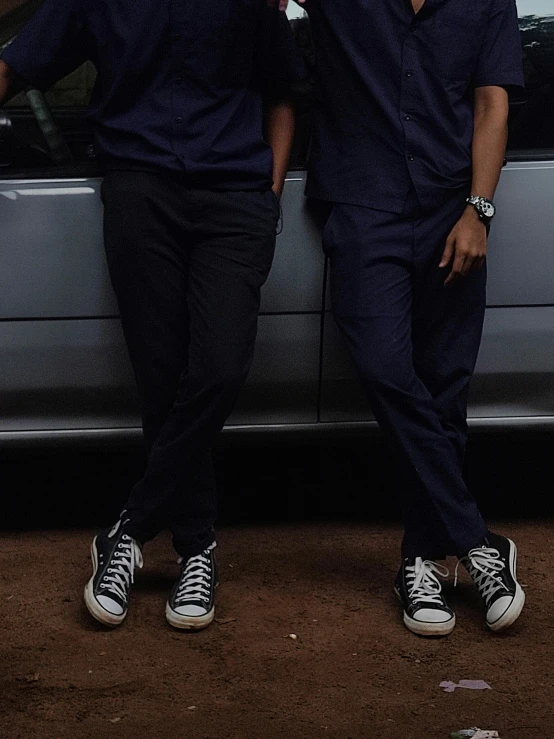 two men wearing black clothes and converse shoes in front of a car