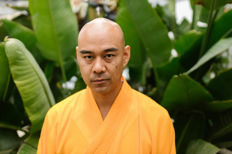 an asian man wearing a bright orange robe standing in front of green plants