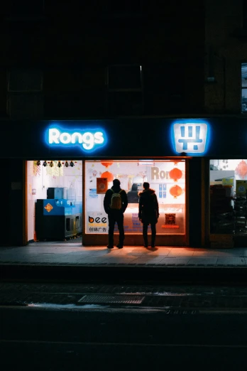 two people standing outside of a store lit up at night