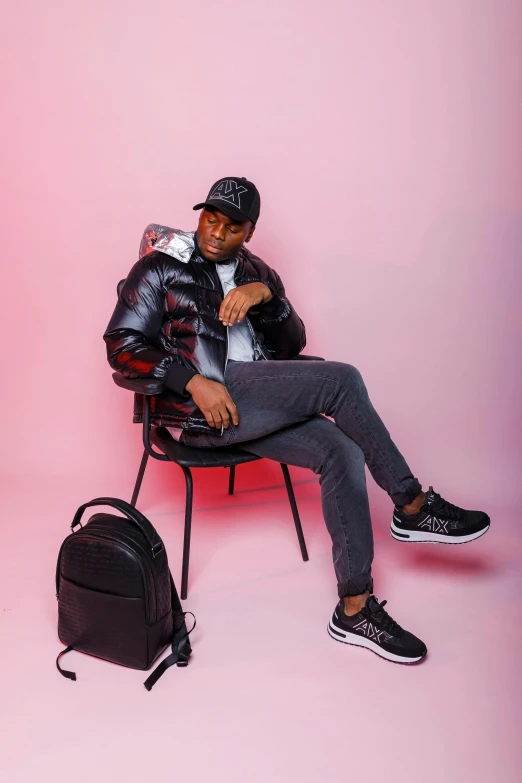 an african american man wearing a black leather jacket and hat while sitting in a chair, with a backpack