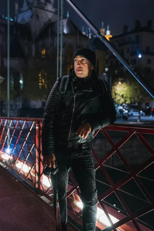 man in a black leather jacket standing on a red bridge at night