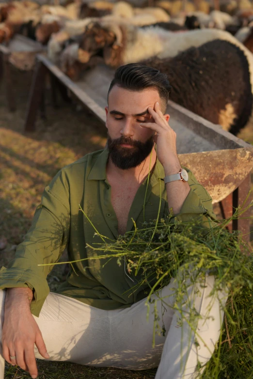 a man sits on a chair and examines the grass on his hands as a flock of sheep stand in the background