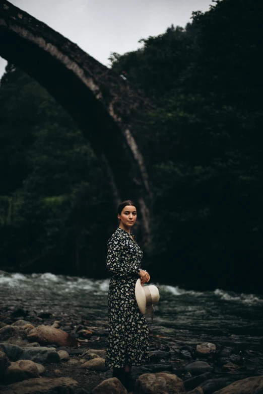 a woman in a long dress and hat stands on rocks near a stream