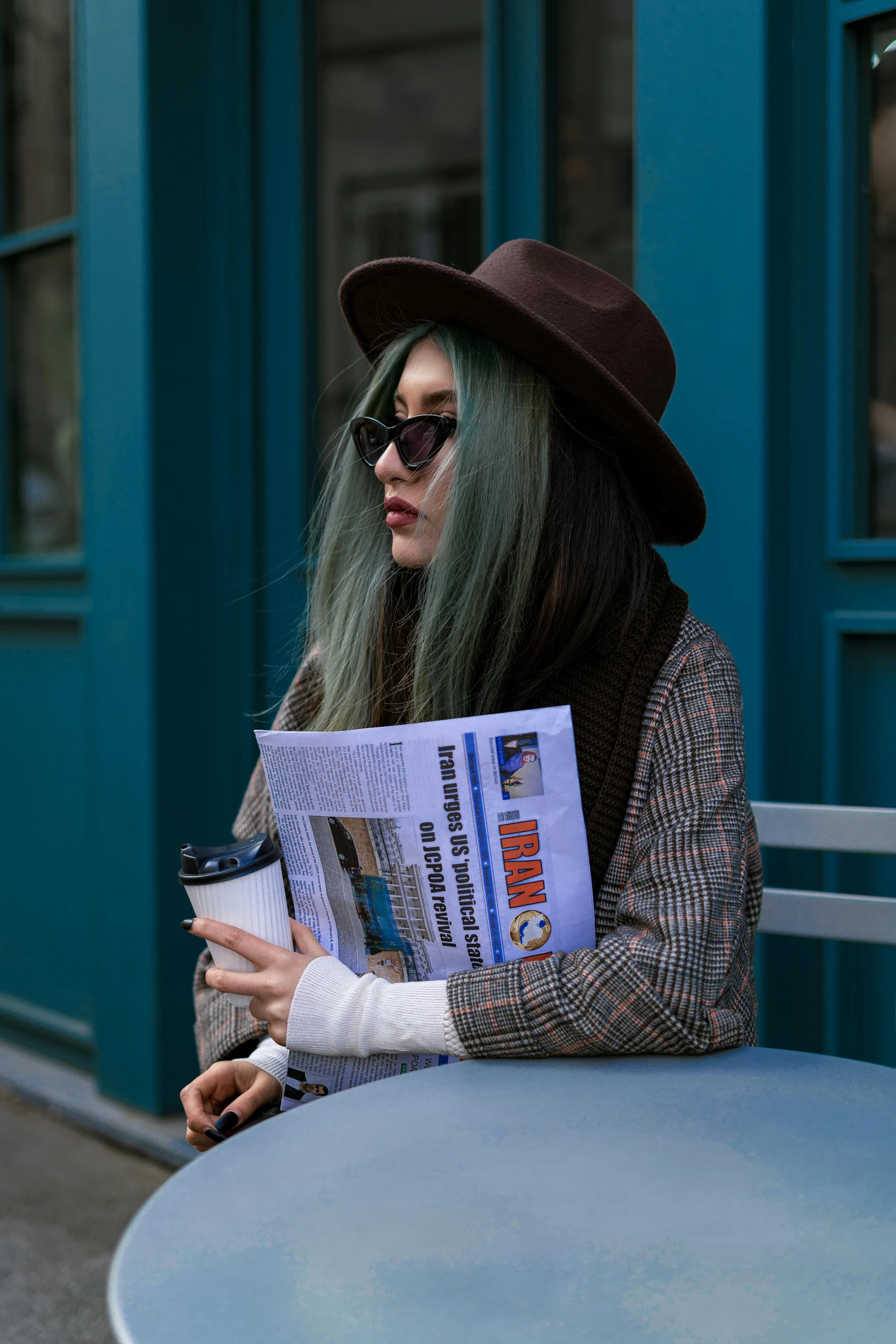 woman with a hat and sunglasses is sitting at a table reading the news