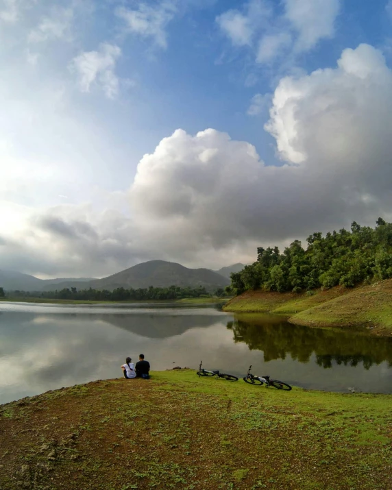 two people sit near their bicycles at the edge of a lake