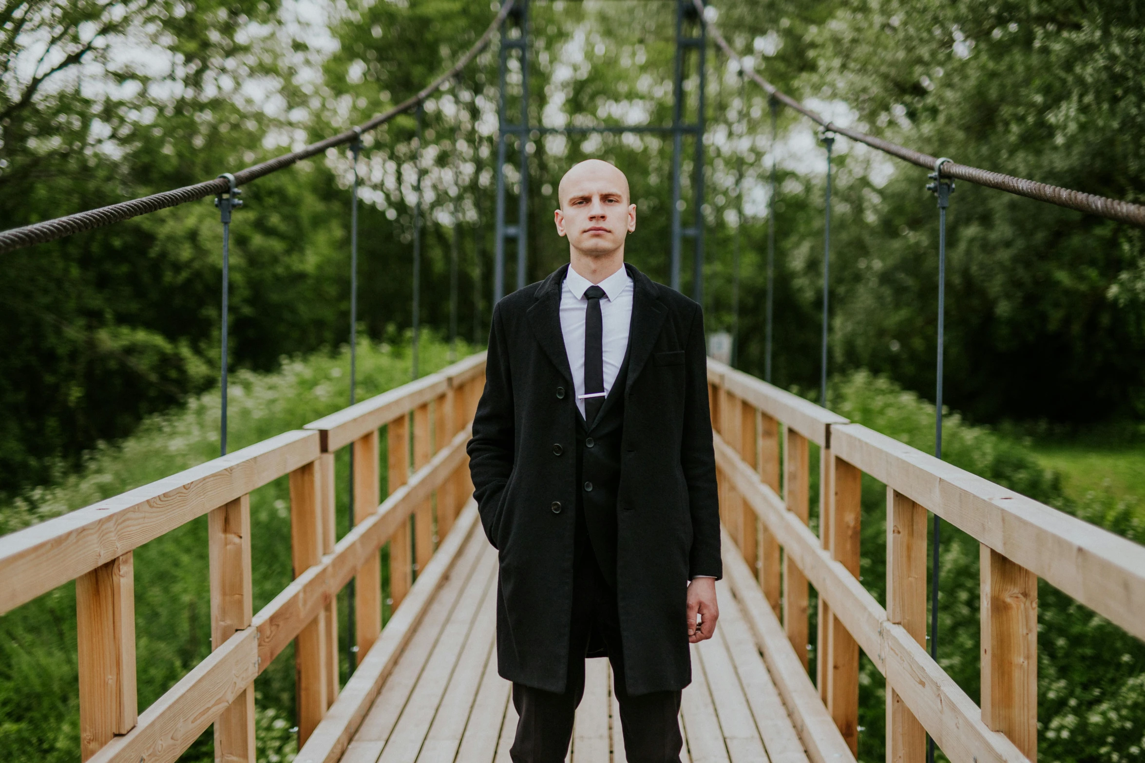 a man in black coat and tie standing on wooden bridge