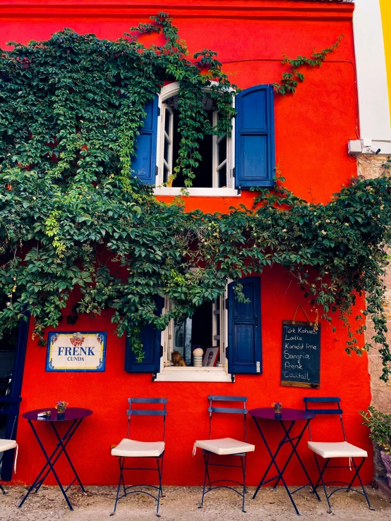 several tables and chairs under a tall red building with a green vine on the wall