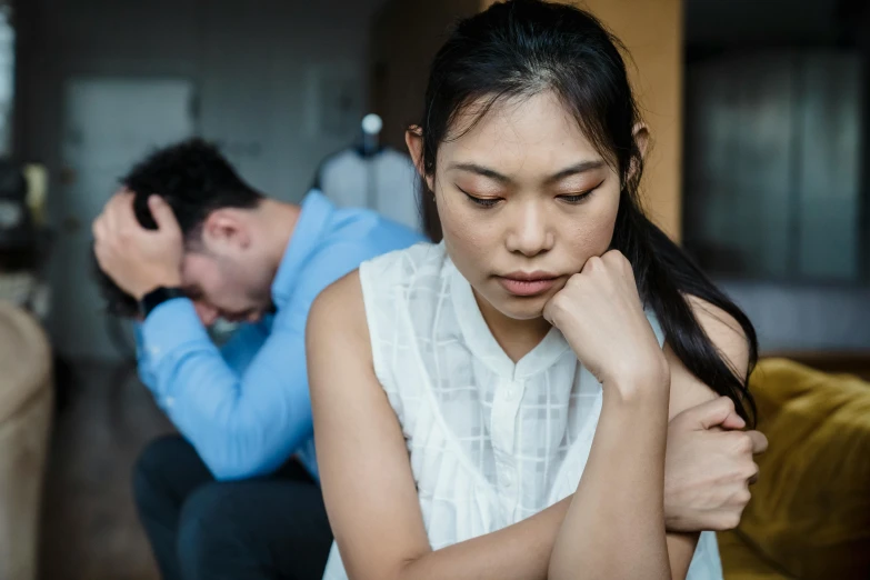 an asian woman wearing white stares away from the camera while a man sits in a chair