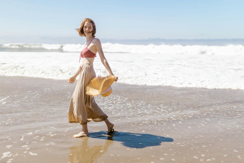 a woman walking across a beach holding a straw hat