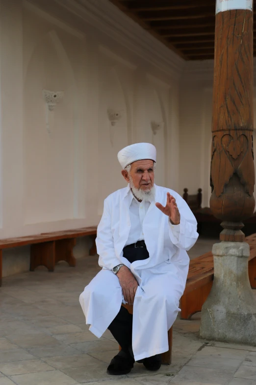 an old man wearing a white outfit seated on a wooden bench