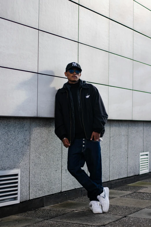 a man posing next to a wall, wearing sunglasses and jeans