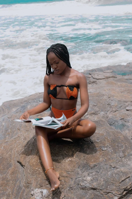 a woman in a bikini sits on the rocks reading a newspaper