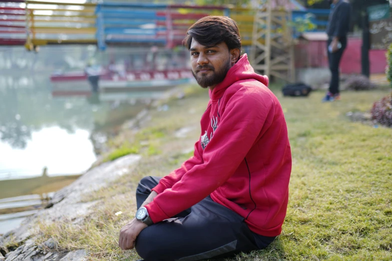 a man sitting on grass near water while another man watches from the other side