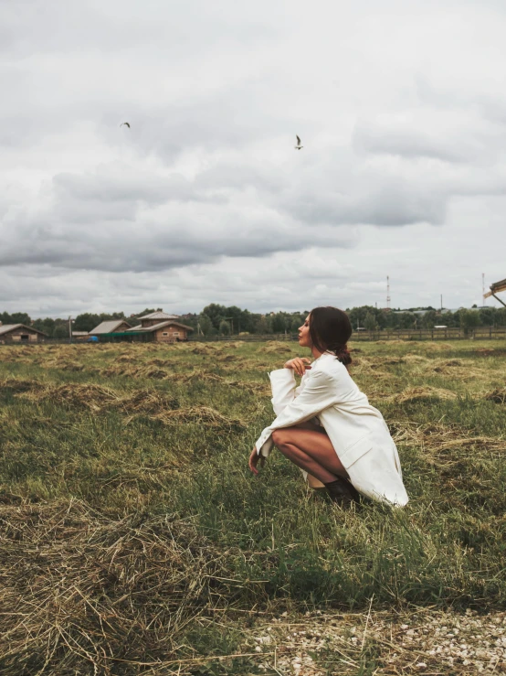 a woman crouches in the grass on the other side of a large field with kites flying high in the sky behind her