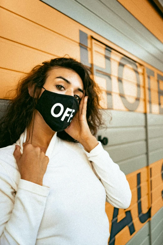 a woman in a white shirt and black mask holding up a cell phone to her ear