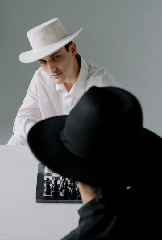 a man wearing a white hat and sitting at a table in front of a chess player
