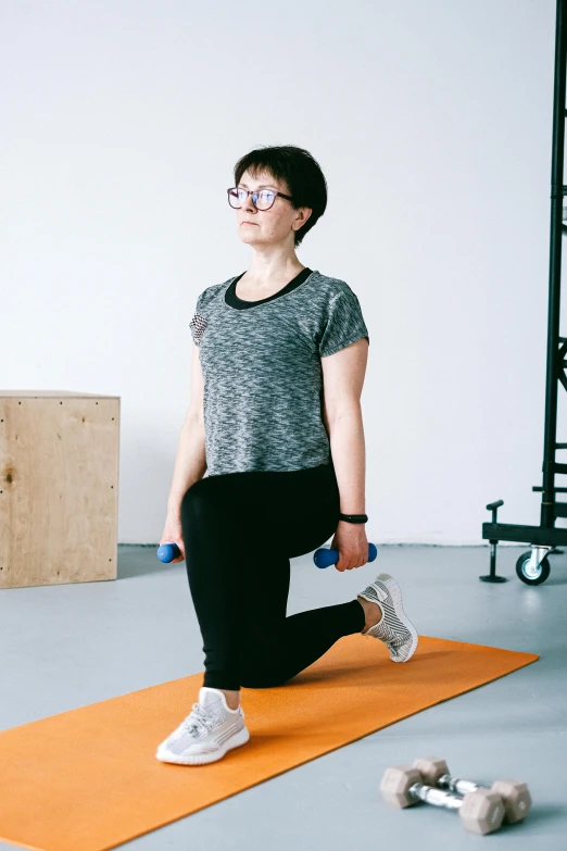 a woman in grey shirt and black pants standing on an orange mat holding two blue dumbs
