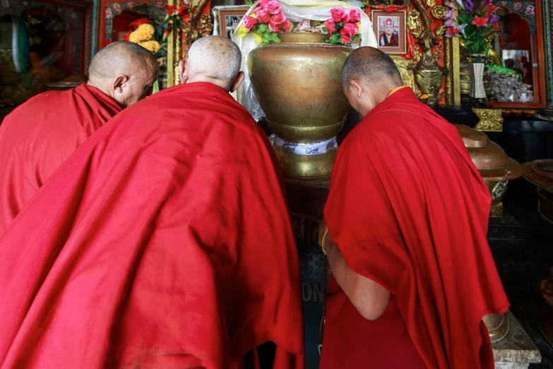 three buddhist monks, each in red robes and headgear, admiring a statue