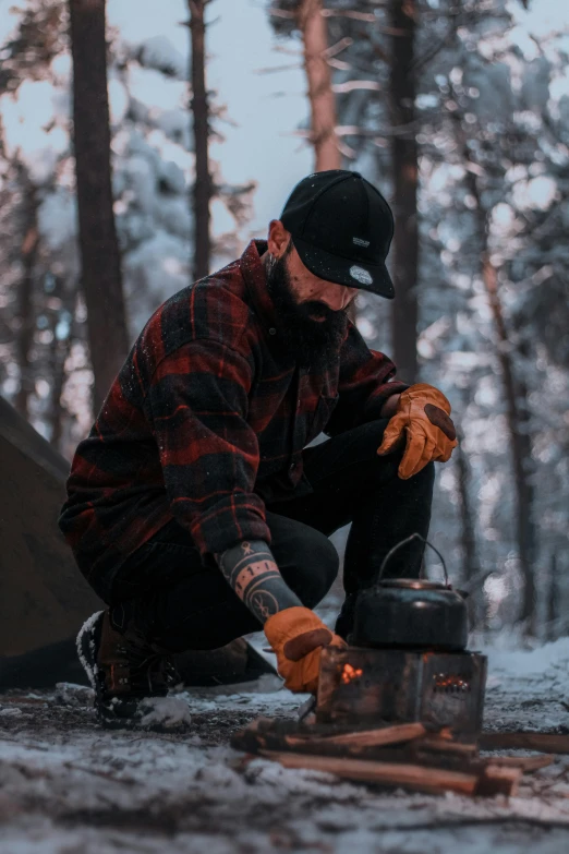 a man with gloves on, sitting down and grilling food on a stove in the snow