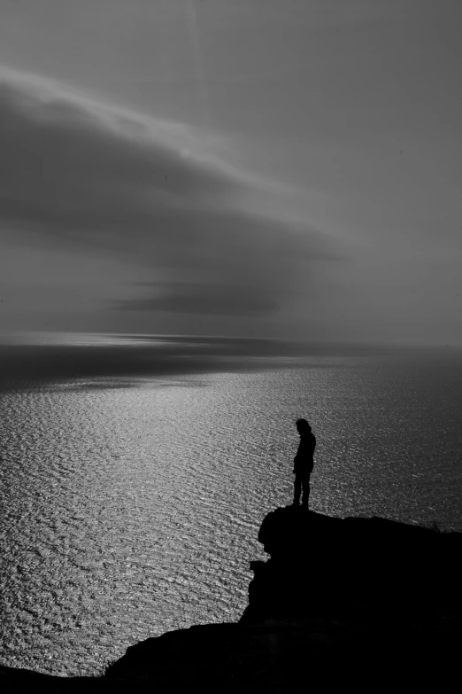 a black and white po of a person standing on top of a cliff by the ocean