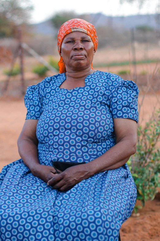 an older woman in a blue dress sits on a dirt path