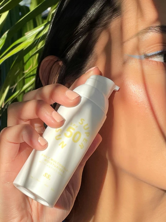 a woman's hand holds a container of sunscreen over her face