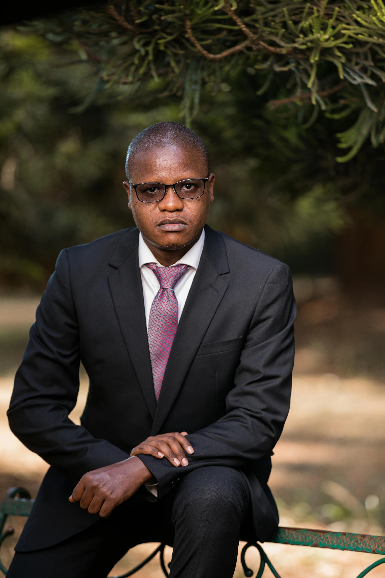 a man in suit and sunglasses sitting on bench under tree