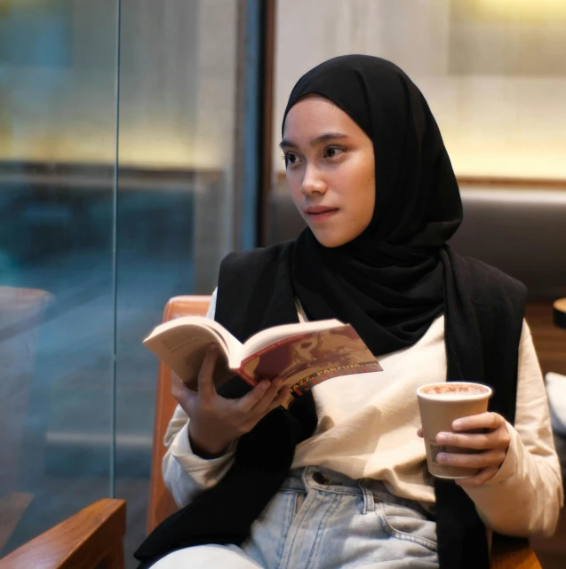 young woman reading a book and drinking coffee while sitting at a table