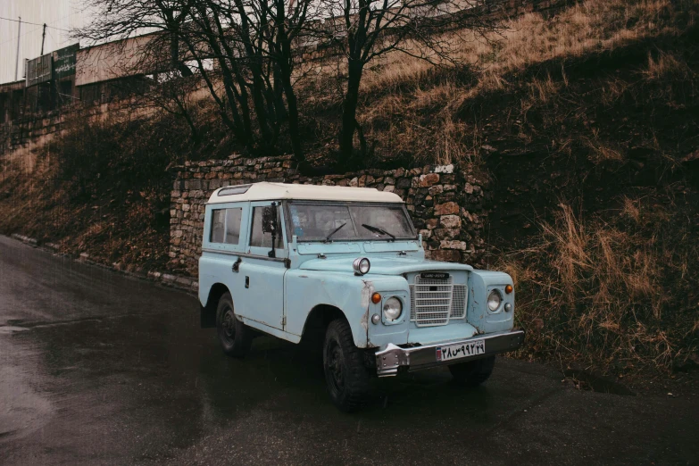 a vintage po of an old, pale blue land rover vehicle parked on a street with brown grass and trees on the side