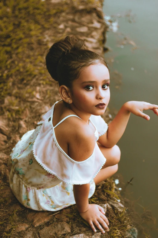 a small girl in a white dress sitting on the side of a river