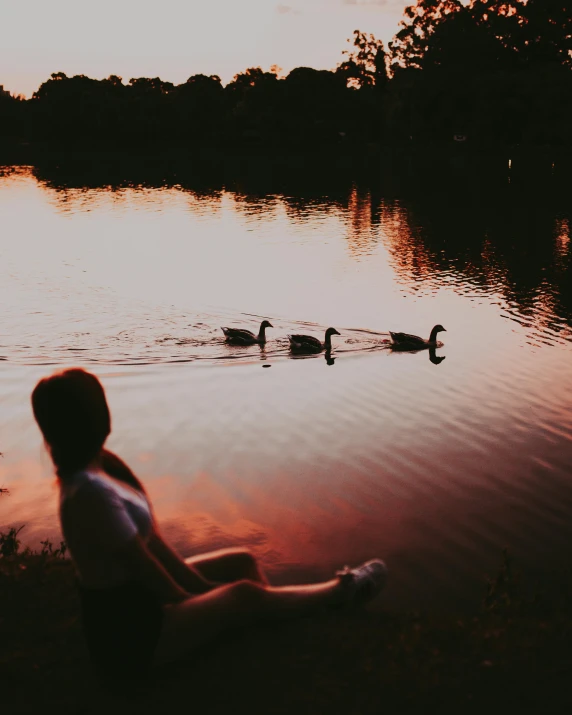 a woman sitting on the bank of a lake watching ducks swim by