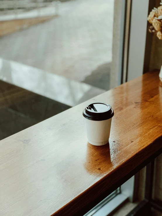 coffee cup with black lid sitting on a window sill by a vase with flowers