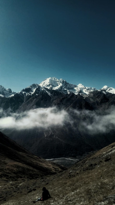 a person sits on the hill overlooking mountains with clouds rolling in