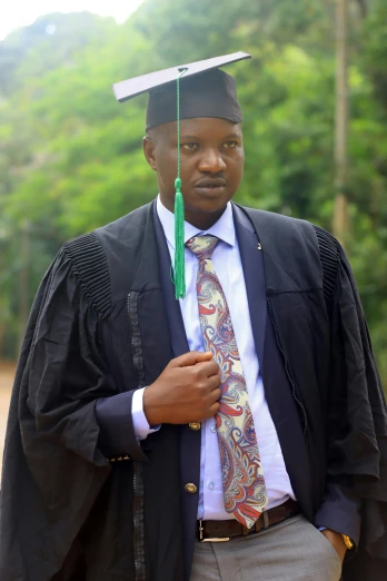a man wearing a black cap and gown standing in front of a green forest