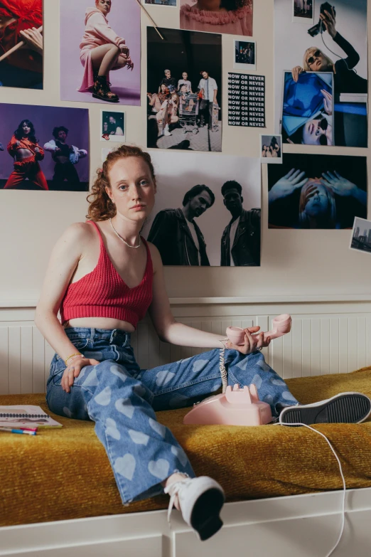 a woman wearing blue jeans sits on a bed in front of a large number of pictures on the wall