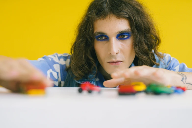 a man wearing a blue shirt with long hair sitting behind a table holding toy cars