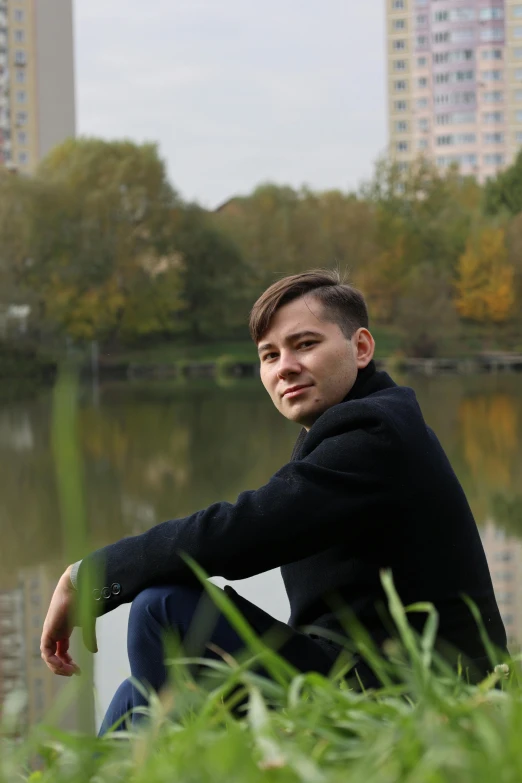 a young man sitting next to a lake with tall buildings