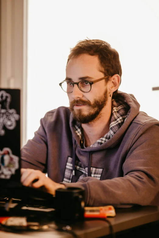 a man wearing glasses and a hoodie sitting in front of a laptop computer