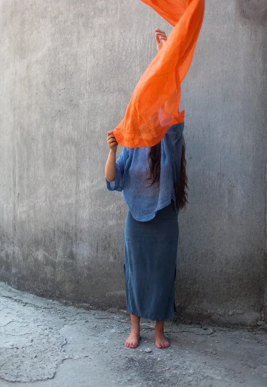 a woman standing against a wall holding an orange scarf over her head