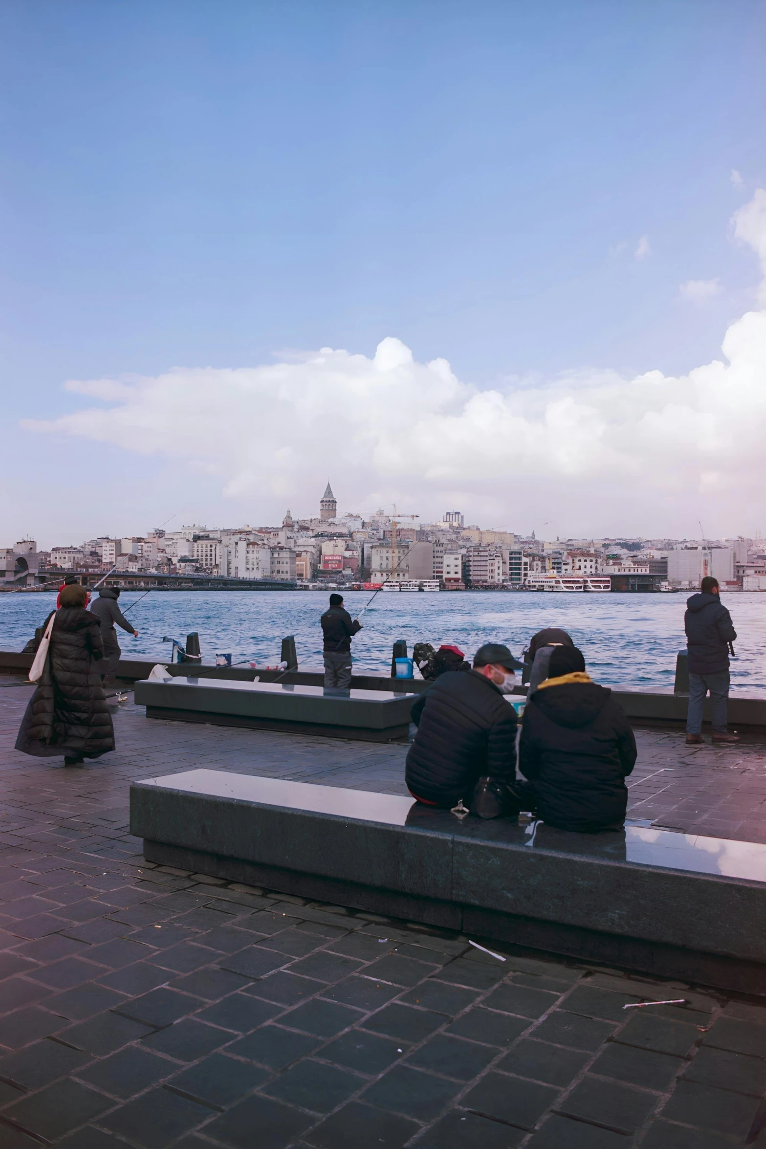 people in coats and black jackets sit on benches next to a body of water